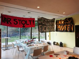 Lokal med bord och stolar i mitten. Längs väggarna hänger banners med queerpolitiska budskap.
