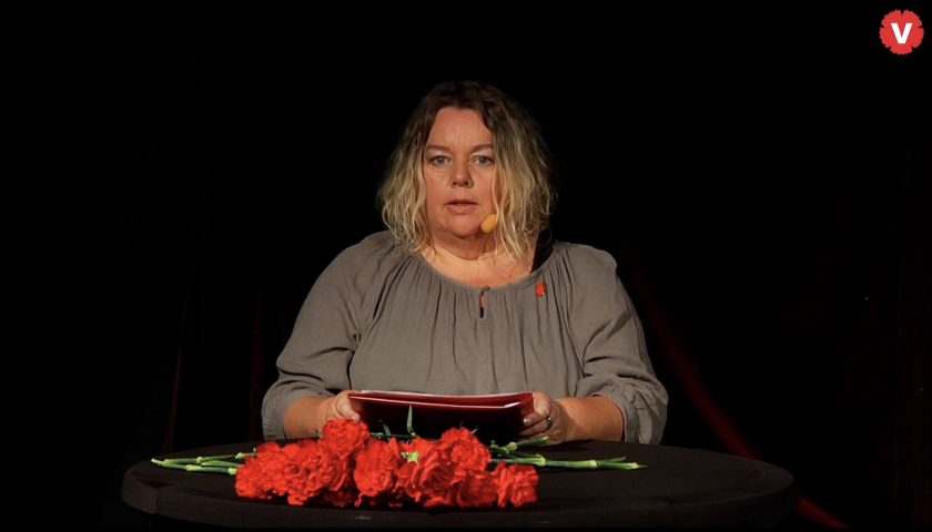 Maria Wåhlin står bakom ett bord med talarkort i händerna. Framför henne på bordet ligger röda nejlikor.